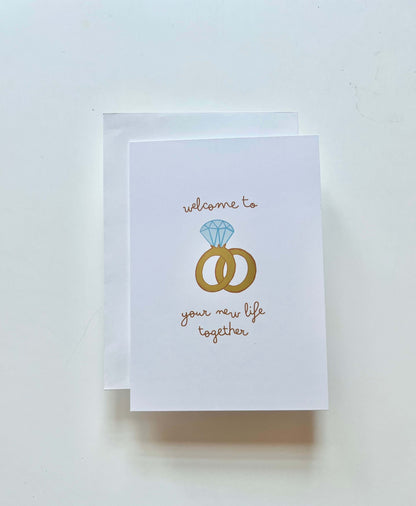 hand drawn greeting card with a diamond ring and wedding band that says welcome to your new life together.