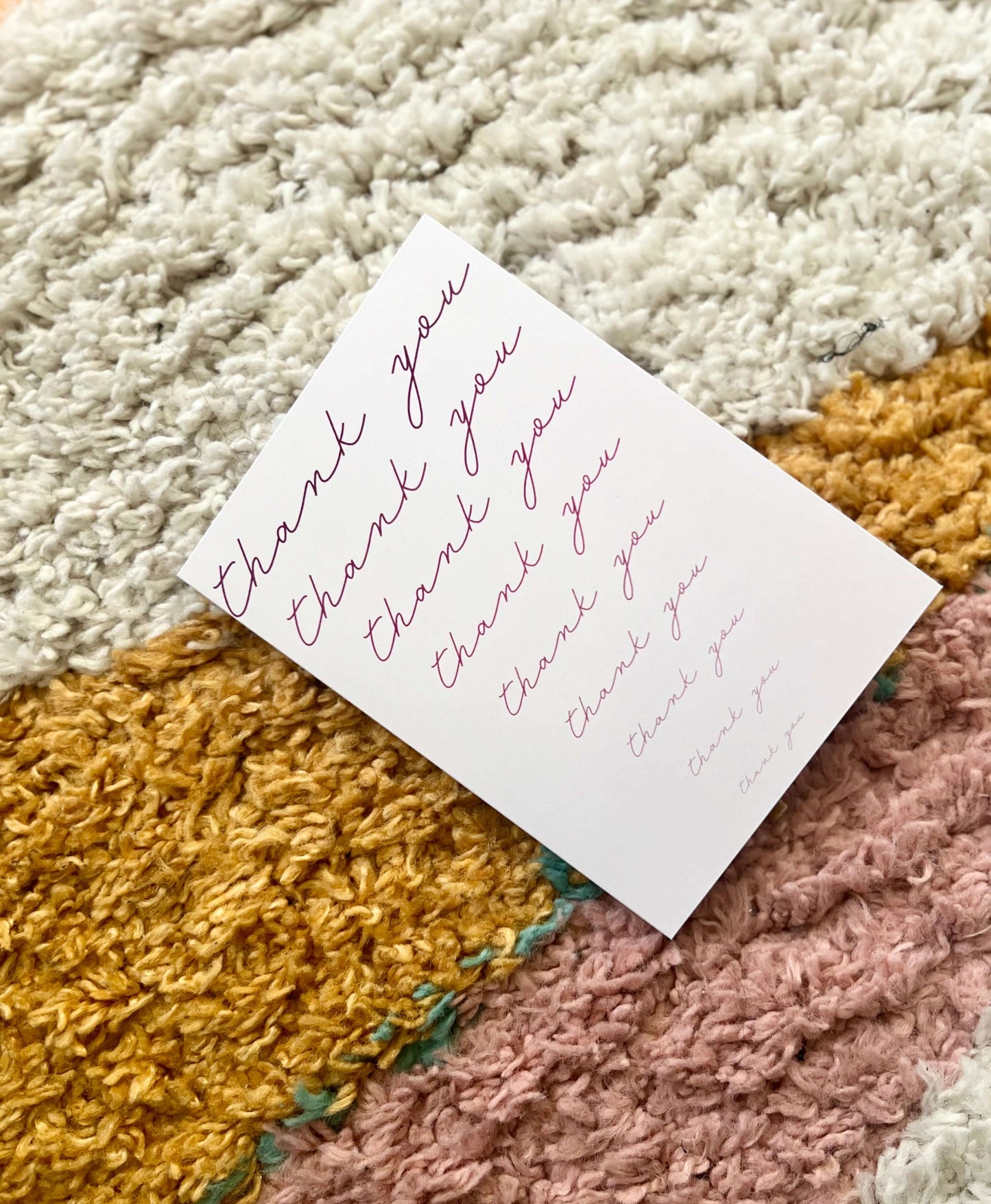 Hand drawn thank you card with cursive text on a pink background, placed on a textured woven rug.