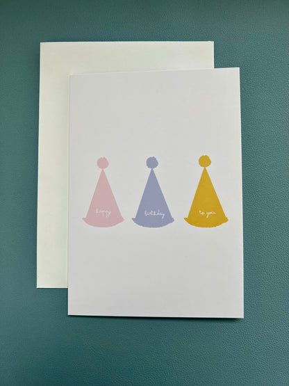 hand drawn greeting card with white background and pink blue and yellow party hats that say happy birthday to you.